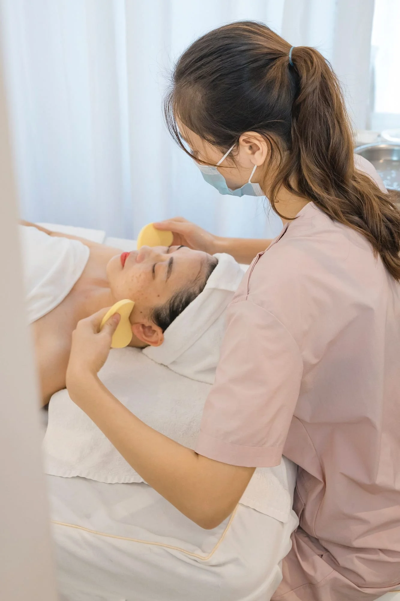 A masked practitioner cleansing a client’s face with a soft sponge.