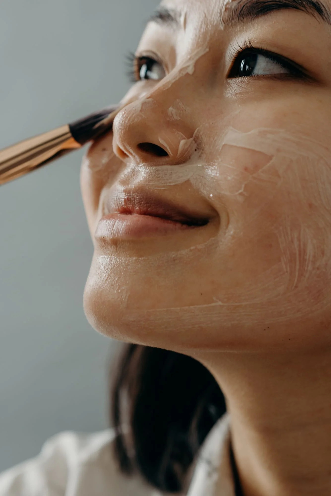 A woman smiling as a brush smooths cream across her cheek.
