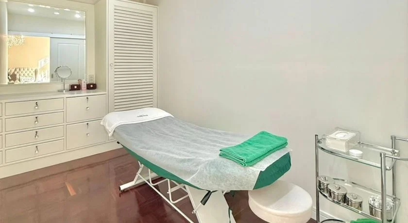 A treatment room: a freshly made bed with a green towel, white drawers and a vanity mirror.